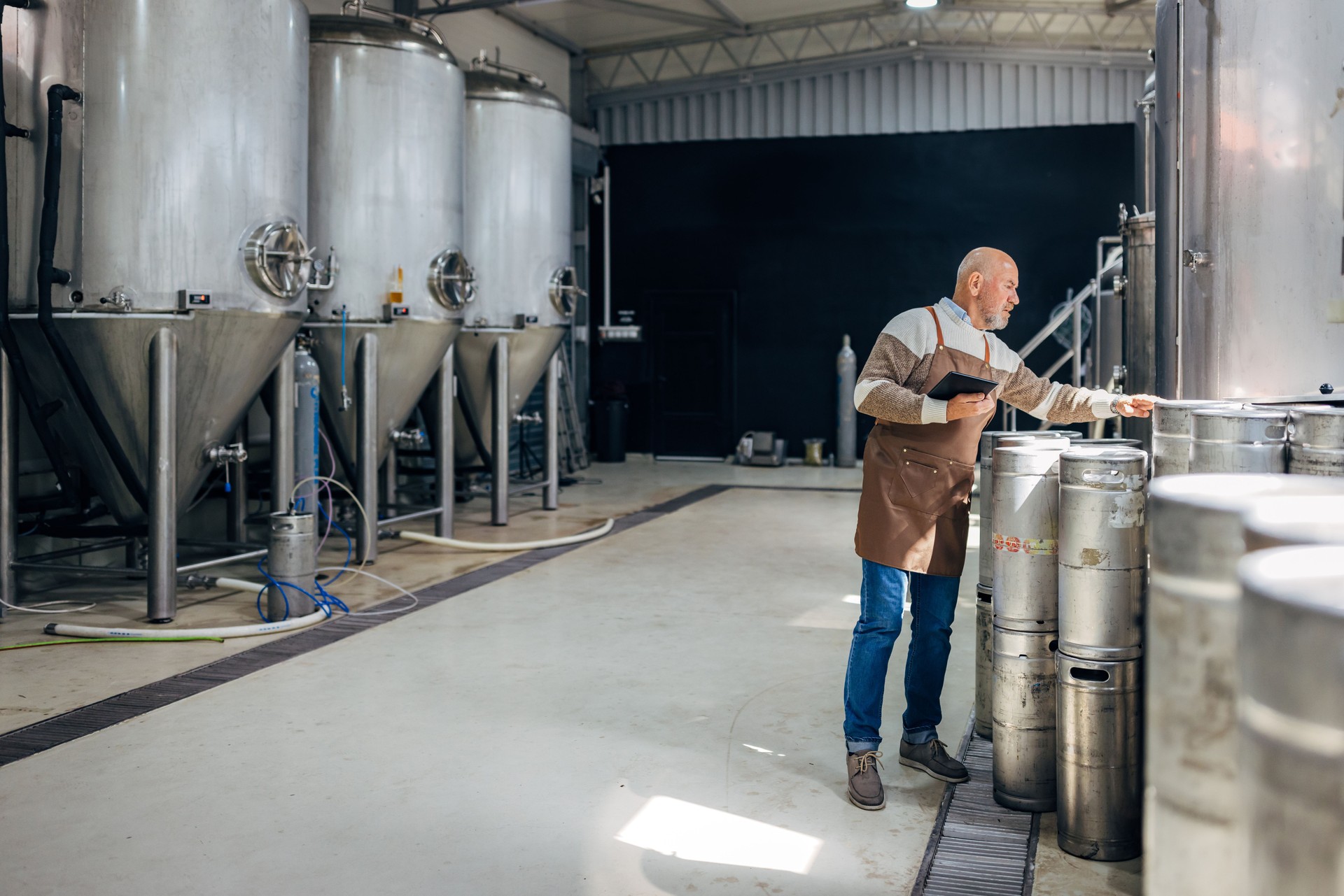 A brewer checks beer barrels in a brewery while using a digital tablet